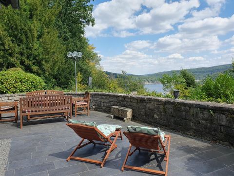 Ein gemütlicher Außenbereich mit Liegestühlen und einer schönen Aussicht auf den Fluss. Im Hintergrund sind Bäume und ein teilweise bewölkter Himmel zu sehen.