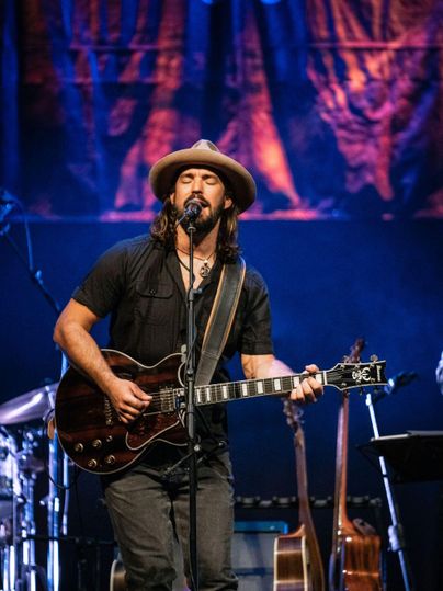 A musician is playing live on stage and singing into a microphone. He is wearing a hat and holding a guitar in his hand.