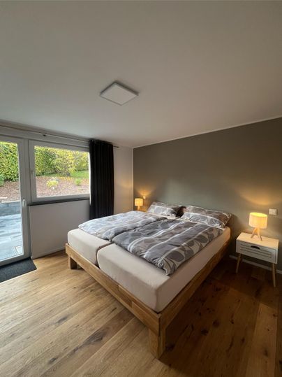 Een gezellige slaapkamer met een groot bed en een houten vloer. Er zijn ramen met uitzicht naar buiten en twee nachtkastjes met lampen.