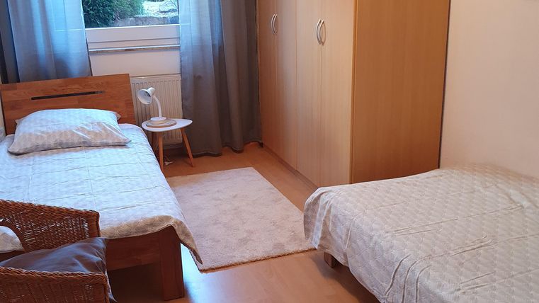 A simple bedroom with two beds and a large wardrobe. There are curtains in front of the window, and a small table stands next to one of the beds.