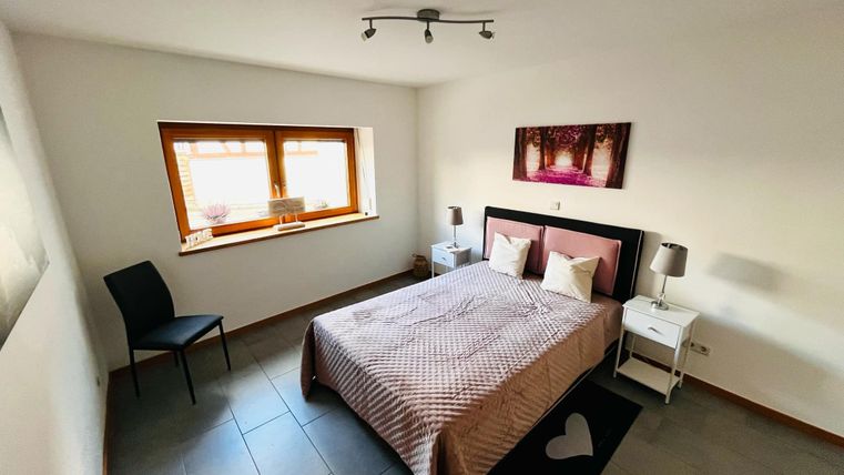 Een gezellig slaapkamer met een tweepersoonsbed, nachtkastjes en een stoel. De muren zijn licht, en een groot raam zorgt voor veel licht.
