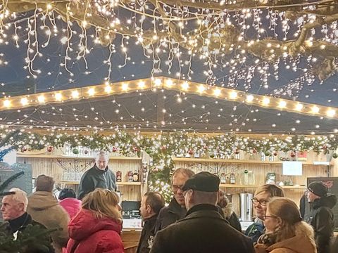 Ein festlicher Weihnachtsmarkt mit vielen Menschen, die sich unter einem beleuchteten Stand versammeln. Lichter und Dekorationen schaffen eine gemütliche Atmosphäre.