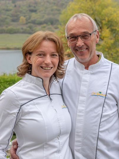 A friendly couple in chef jackets is smiling in front of a picturesque landscape. In the background, trees and a body of water can be seen.