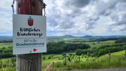 A guidepost for hikes in nature shows a beautiful landscape with gentle hills and wooded areas. The sky is cloudy, giving the scene a calm atmosphere.