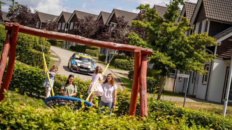 Un terrain de jeu avec des balançoires et une voiture colorée en arrière-plan. Quelques enfants jouent joyeusement en plein air.