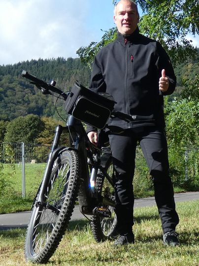 Ein Mann steht neben seinem Mountainbike in einer grünen Landschaft. Er lächelt und zeigt Daumen hoch.