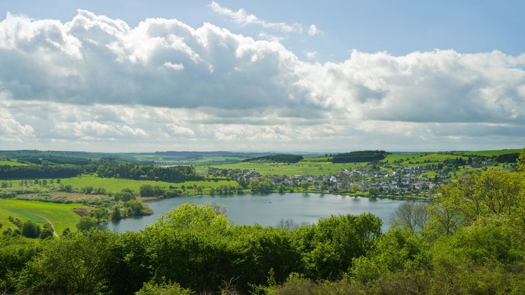 Landschaft mit See, grünen Feldern und Dorf im Hintergrund unter bewölktem Himmel.