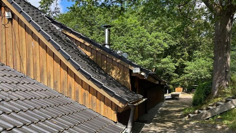 A rustic house with a sloping, paved roof and wood siding. In the background, trees and a narrow path are visible.