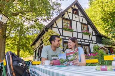 Zwei Personen sitzen draußen in einer Gastronomie und sehen sich einen Flyer an. Vor ihnen stehen zwei Kaffeetassen und im Hintergrund sieht man das Café, welches ein altes Fachwerkhaus ist.