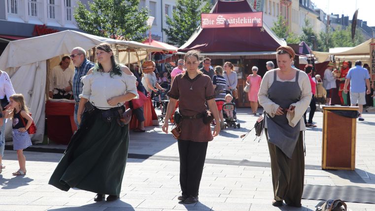 Drie vrouwen in historische kleding lopen over een marktplein. Op de achtergrond zijn marktkraampjes en veel mensen te zien.