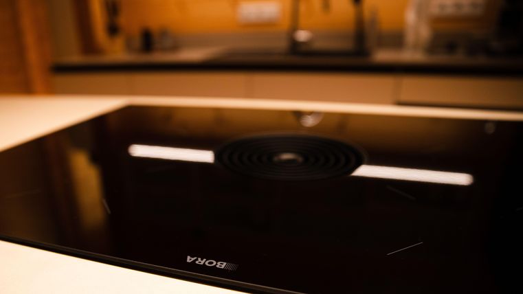 Ein modernes Kochfeld mit einer eleganten schwarzen Oberfläche. Im Hintergrund ist eine stilvolle Küche mit Holz- und Grautönen sichtbar.