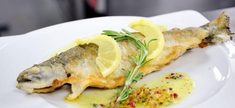 Un poisson grillé dans une assiette, garni de tranches de citron et de romarin. Il est accompagné d'une sauce savoureuse.