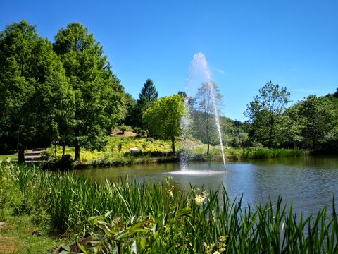 Een vijver in de spa-tuinen met een waterfontein, omringd door groene bomen en planten onder een strakblauwe hemel.