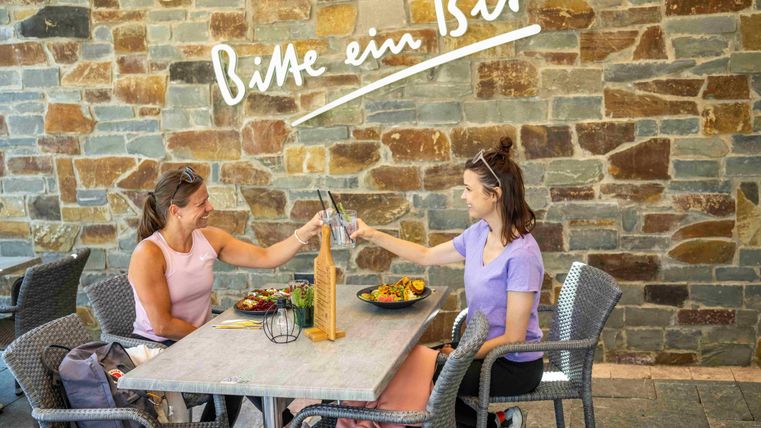 Zwei Frauen stoßen mit Gläsern an einem Tisch an. Im Hintergrund ist eine Steinmauer mit der Aufschrift „Bitte ein Bit“ zu sehen.