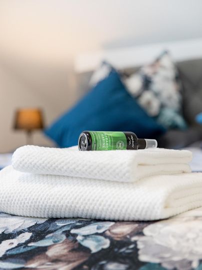 Op een bed liggen twee gevouwen witte handdoeken. Daarop staat een fles met groene inhoud en een decoratief kussen op de achtergrond.