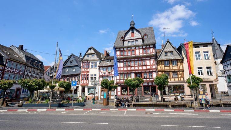 Eine malerische Straße mit historischen Fachwerkhäusern und bunten Fahnen. Der Himmel ist blau und der Tag sonnig.