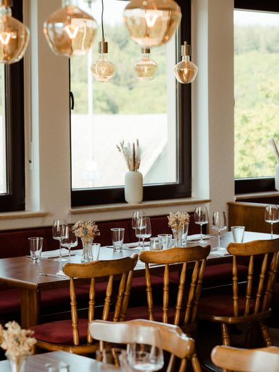 Ein gemütliches Restaurant mit Holzstühlen und elegantem Tischgedeck. Große Fenster lassen viel Licht herein und bieten einen Blick auf die grüne Landschaft.