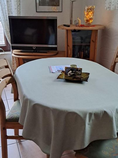 A dining room with a round table set with a light tablecloth. In the background, there is a television and a fireplace, surrounded by a cozy atmosphere.