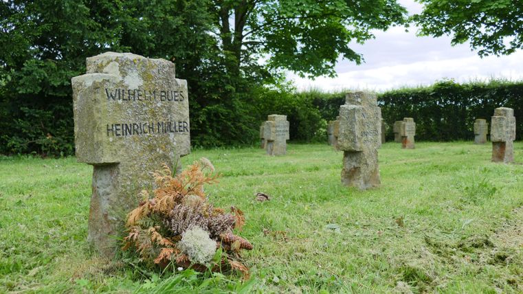 Een begraafplaats met verschillende stenen grafstenen, gelegen op een groene weide. Op sommige grafstenen zijn namen gegraveerd en op een van de graven ligt een klein boeket bloemen.