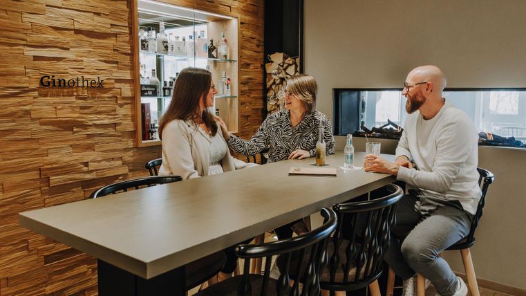 Een gezellige bar met houten decor, waar drie personen aan een tafel zitten. Ze hebben een levendig gesprek en genieten van de sfeer.
