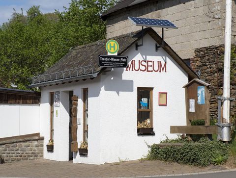 Ein kleines Museum mit weißer Fassade und einem Sonnensystem auf dem Dach. Es liegt an einer Straße mit einem Haltestellenschild in der Nähe.