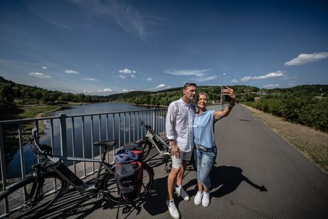 Un couple prend un selfie sur un pont au bord du lac de Kronenburg, avec des vélos en arrière-plan.