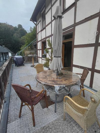A terrace with a round wooden table and several chairs. In the background, a traditional half-timbered house can be seen.