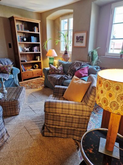 A cozy living room with multiple seating options. There are bookshelves, plants, and warm light from a lamp.