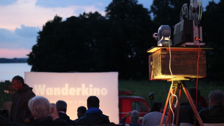 An outdoor movie night with people watching a film. In the foreground is a projector projecting an image onto a screen.