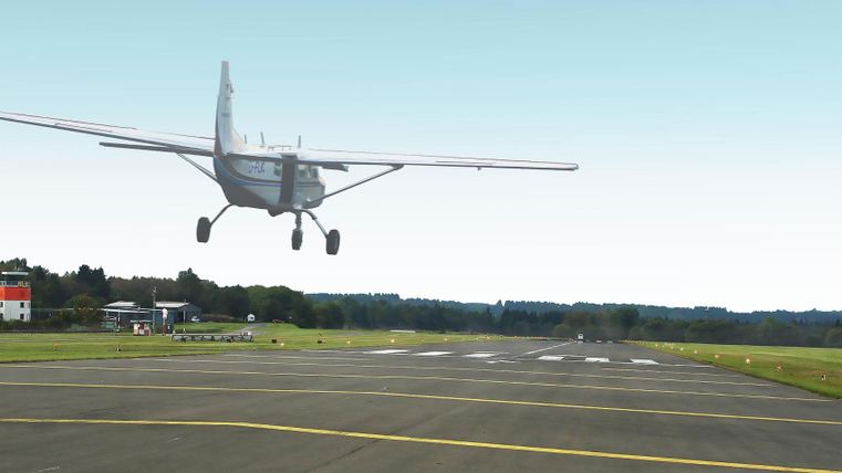 A small airplane is landing at an airport. In the background, green fields and a building can be seen.