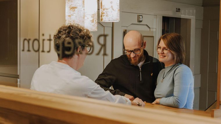 Een paar staat bij de receptie en spreekt met een medewerkster. Op de achtergrond zijn decoratieve lampen en een spiegel te zien.
