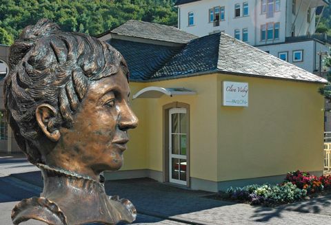 Eine Bronze-Büste einer Frau steht vor einem gelben Gebäude. Im Hintergrund sind Bäume und weitere Gebäude zu sehen.