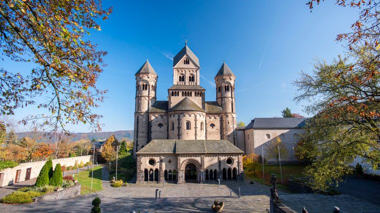 Eine imposante Kirche mit Türmen und einem klaren blauen Himmel. Im Vordergrund sieht man einen Platz, umgeben von Bäumen.