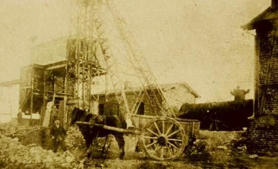 Une vieille image d'un chantier avec une locomotive à vapeur et une structure de levage. Un cheval tire une charge de wagon près du lieu de travail.