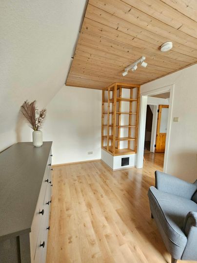 Een heldere, uitnodigende ruimte met houten vloer en open plafond. In de hoek staat een boekenplank, en een comfortabele stoel completeert de inrichting.