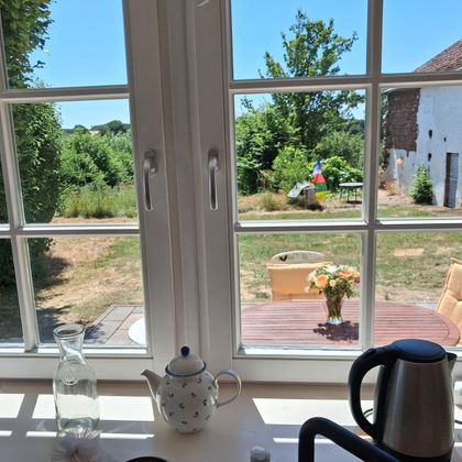 Un regard par la fenêtre sur un jardin par temps ensoleillé. Au premier plan, on peut voir quelques tasses à café et une cafetière.