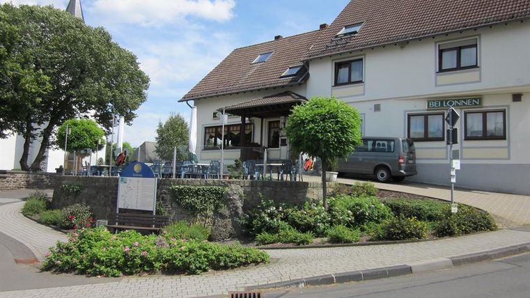 Blick von vorne auf das Gasthaus Bei Lonnen.Das zweistöckige Gebäude ist hell angestrichen und hat ein schwarzgedecktes Dach. Vor der Gäststätte sieht man eine runde Terrasse, die mit mehreren Tischen und Stühlen ausgestattet ist.