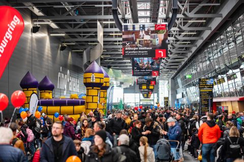 Een grote menigte in een hal met een grote, kleurrijke springkussen op de achtergrond. De sfeer is levendig met veel gezinnen en kinderen.