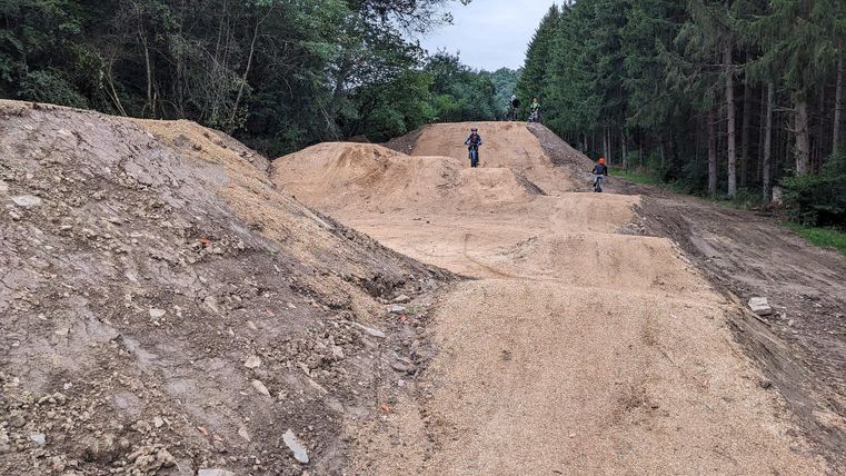 Eine Mountainbike-Strecke mit Hügeln und Sprüngen im Wald. Im Hintergrund sind zwei Biker zu sehen.
