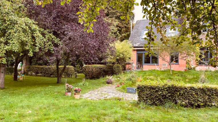 Un beau jardin avec différents arbres et une pelouse bien entretenue. À l'arrière-plan, on peut voir une maison rouge avec des fenêtres.