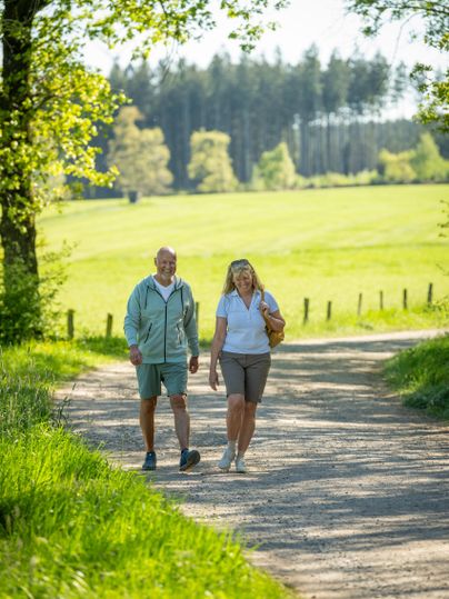 Un couple âgé se promène sur un chemin tranquille à travers un paysage verdoyant. Le soleil brille et les arbres offrent de l'ombre.