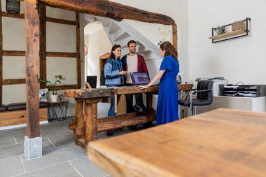 Helle Hotelrezeption mit rustikalen Holzbalken, an der eine Mitarbeiterin in blauem Kleid mit zwei Gästen am Tresen spricht. Im Hintergrund stehen Treppe, Laptop, Drucker und Ablagen, die den persönlichen Empfangsbereich des Hotels zeigen.