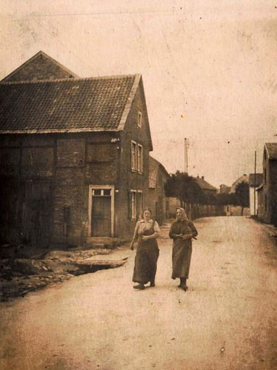 Two women are walking down a quiet street lined with old buildings. The scene has a nostalgic feel and looks historic.