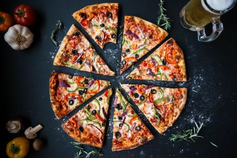 Een vers gebakken pizza, in stukken gesneden, ligt op een donkere tafel. Naast de pizza staat een glas bier en enkele verse ingrediënten.