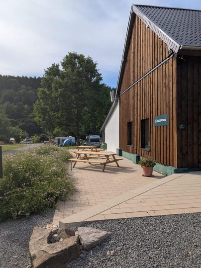 Une maison en bois moderne avec une terrasse et une table en extérieur. En arrière-plan, des arbres et des terrains de camping sont visibles.