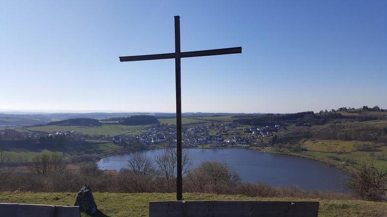 A large cross stands on a hill overlooking a lake and surrounding green landscapes. The clear sky and calm scenery evoke a peaceful atmosphere.