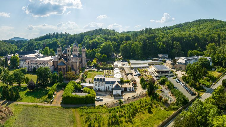 Luftaufnahme der Abtei Maria Laach, eingebettet in eine grüne Hügellandschaft; die romanische Klosteranlage mit ihrer markanten Kirche und den umliegenden Gebäuden liegt ruhig und sonnig zwischen dichten Wäldern.