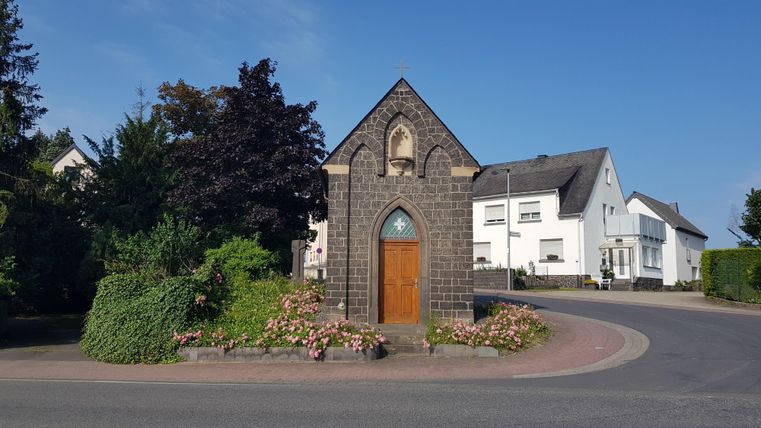 A small chapel made of dark stone stands at a bend in the road. Green plants and flowers frame the entrance.