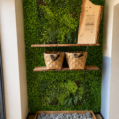 Een groene wand met planten en twee houten planken. Op de planken staan gevlochten manden en een houten bord met een inscriptie.