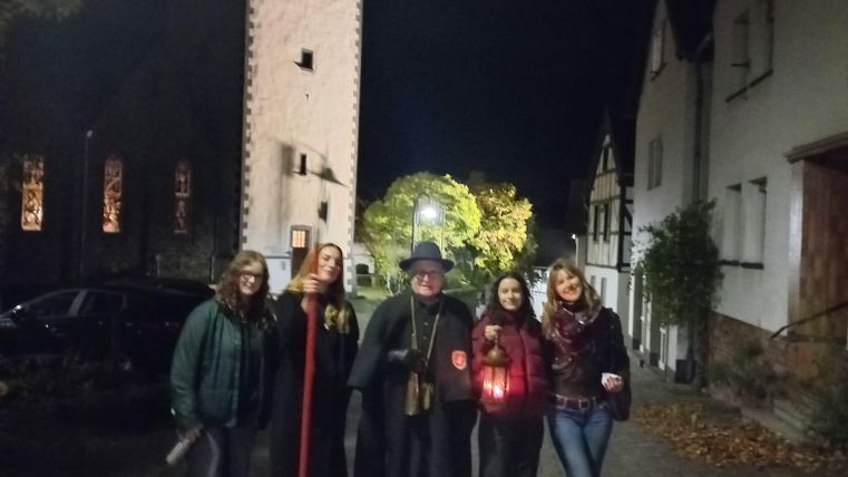 Eine Gruppe von Menschen steht im Dunkeln um einen Baumstamm und hält Fackeln. Eine Person in einem langen Mantel spricht zu ihnen, während sie aufmerksam zuhören.